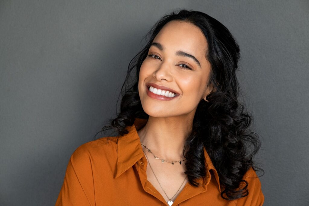 Woman with long black hair in orange shirt smiling with gray background