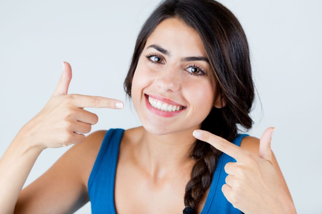 Woman with dark hair in blue shirt pointing to her smile with both hands